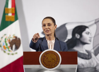 President Claudia Sheinbaum at her daily press conference, standing at the presidential podium in the National Palace in Mexico City. She points at a reporter off camera to take their question.