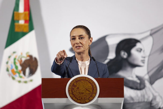 President Claudia Sheinbaum at her daily press conference, standing at the presidential podium in the National Palace in Mexico City. She points at a reporter off camera to take their question.