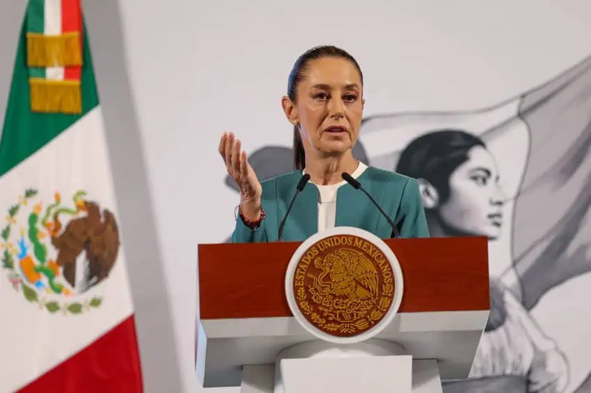 Mexico's President Claudia Sheinbaum at the presidential podium in the National Palace. She is talking to reporters in the press briefing room and holding her right hand up, palm upward in a gesture