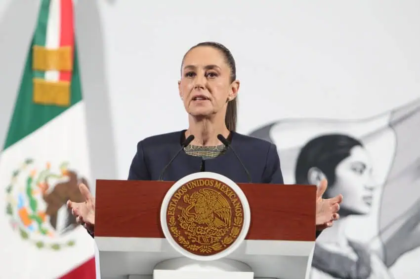 President Sheinbaum during a daily press conference in the National Palace. She is holding her arms in front of her on either side as she speaks to reporters.