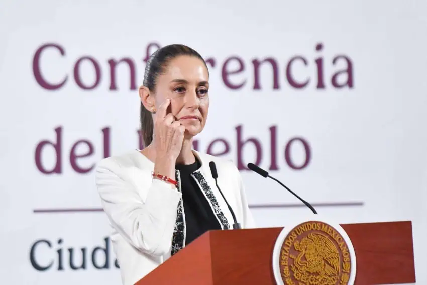 Presidenta Claudia Sheinbaum en su conferencia de prensa diaria. Está parada en el podio presidencial con su dedo índice derecho tocando su mejilla mientras escucha la pregunta de un periodista.