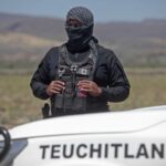 A masked security agent in black clothing holds binoculars while standing behind a vehicle printed with the word Teuchitlán