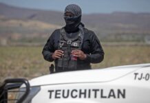 A masked security agent in black clothing holds binoculars while standing behind a vehicle printed with the word Teuchitlán