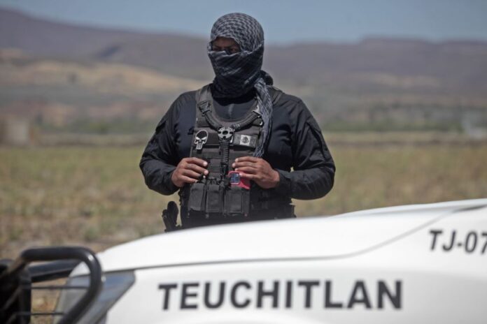 A masked security agent in black clothing holds binoculars while standing behind a vehicle printed with the word Teuchitlán