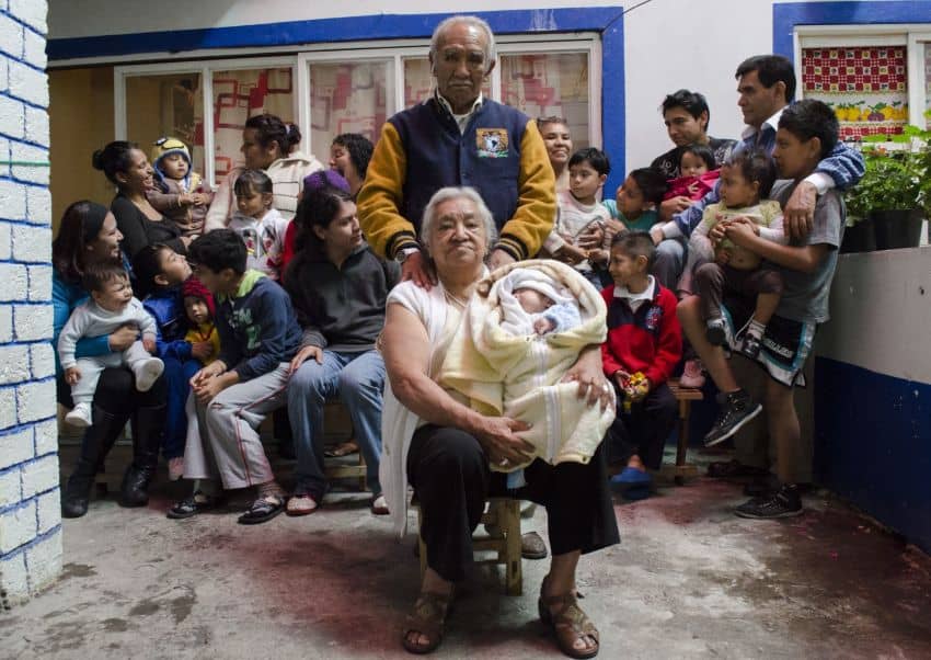 An extended Mexican family posing together, with many men and women holding infants and toddlers and young children sitting among them. At the head in the center is an elderly man and woman, presumably husband and wife. He is standing behind her with his hands on her shoulders. She is seated and holding an infant swaddled in blankets as they both pose for the photo.