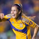 A woman with a long ponytail and wide smile runs past the camera wearing a blue and yellow soccer jersey