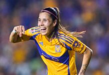 A woman with a long ponytail and wide smile runs past the camera wearing a blue and yellow soccer jersey