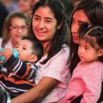 Two young Mexican women sitting in a crowd in a stadium or hall, each with a child on their lap. One of the women is smiling as she talks to the woman next to her.