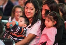 Two young Mexican women sitting in a crowd in a stadium or hall, each with a child on their lap. One of the women is smiling as she talks to the woman next to her.