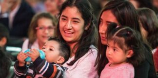 Two young Mexican women sitting in a crowd in a stadium or hall, each with a child on their lap. One of the women is smiling as she talks to the woman next to her.