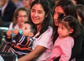 Two young Mexican women sitting in a crowd in a stadium or hall, each with a child on their lap. One of the women is smiling as she talks to the woman next to her.