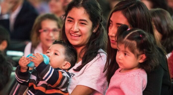 Two young Mexican women sitting in a crowd in a stadium or hall, each with a child on their lap. One of the women is smiling as she talks to the woman next to her.