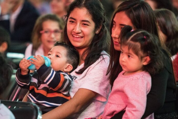 Two young Mexican women sitting in a crowd in a stadium or hall, each with a child on their lap. One of the women is smiling as she talks to the woman next to her.