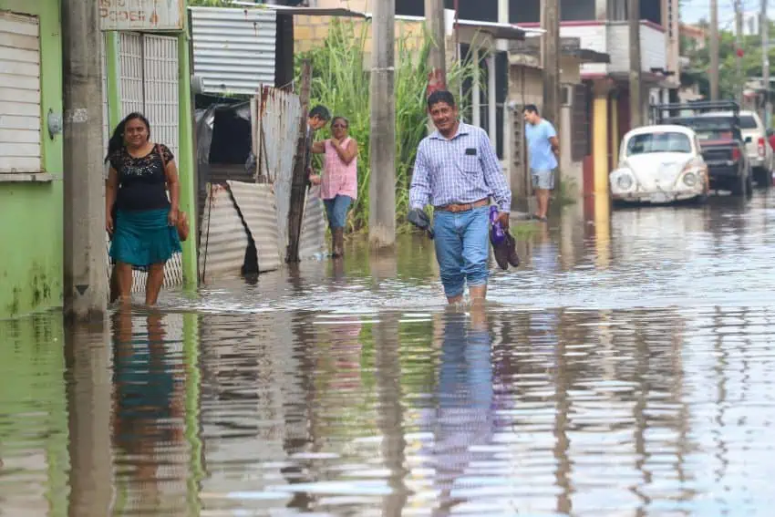 Mexican people walking nonchalantly through flooded streets up to their knees in Villahermosa, Tabasco. One man is walking holding his shoes in his hand and looking at the camera. To the side, a woman walks through the water, also gazing forward.