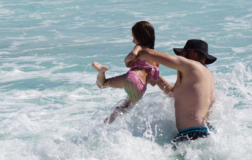 A man holding a little girl over crashing waves in Cancun. She's wearing a pink and white two piece bathing suit and the man is wearing a black bucket hat and black swim trunks with blue stripes at the waist.