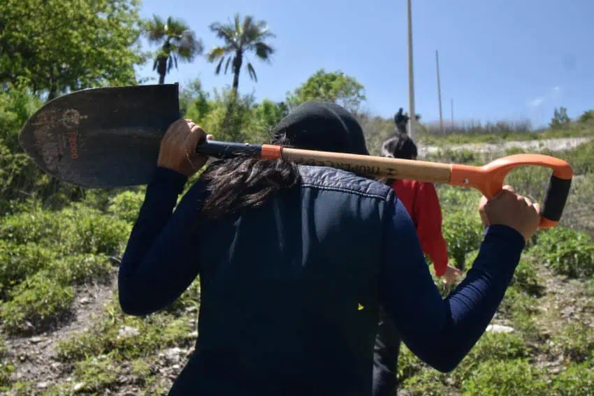 Woman in dark clothing and a baseball cap holds a shovel behind her shoulders as she walks through scrub land. Her back is to the camera.