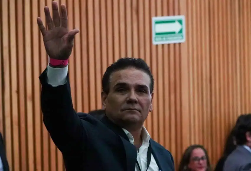 Mexican middle-aged man standing and waving to people off camera. In the background are people sitting on the ground watching him