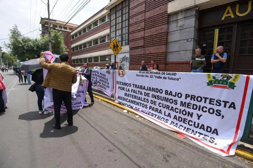 Mexico's Ministry of Health medical workers protesting lack of medicine and medical equipment in Toluca, Mexico state. Protesters stand in front of a large banner on the sidewalk explaining why they are protesting.