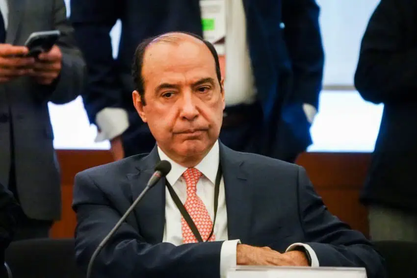 A balding man in a navy men's suit, white shirt and red patterned tie, sitting at a table with a microphone. He looks deep in thought.