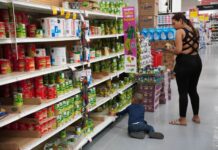 Woman in a Mexican supermarket holding cans of food in her hands while a little boy with her sits on the floor next to her and grabs a can on the lowest shelf.