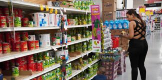 Woman in a Mexican supermarket holding cans of food in her hands while a little boy with her sits on the floor next to her and grabs a can on the lowest shelf.