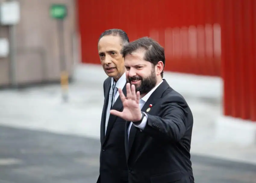 Chilean President Gabriel Boric with an unknown older man. Boric is waving at people off camera. Both men are wearing formal suits.