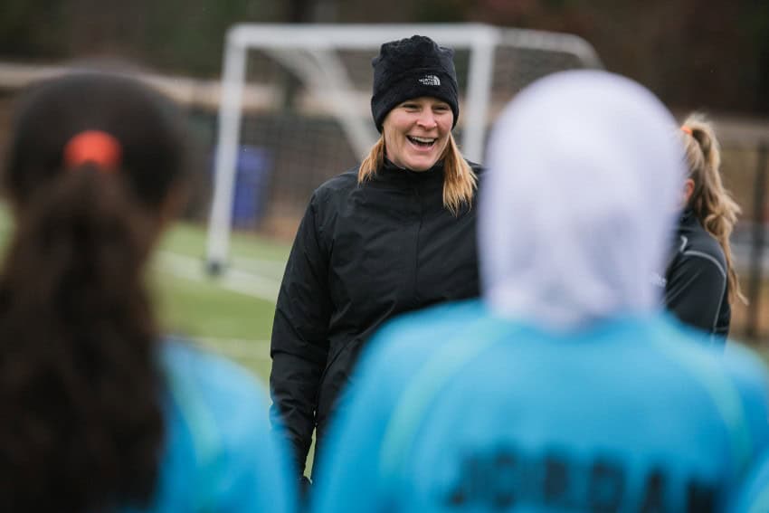 Una mujer con una chaqueta de parka y un sombrero de invierno negro de la cara norte se encuentra en un campo de fútbol y se ríe, ya que está rodeada de mujeres más jóvenes con equipo deportivo para el equipo nacional de fútbol femenino Jordan.