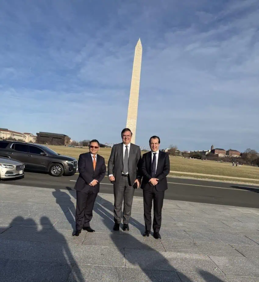 Three men in dark suits smiling for the camera on the side of a roadway with cars. Further in the background of the photo is the Washington Monument.