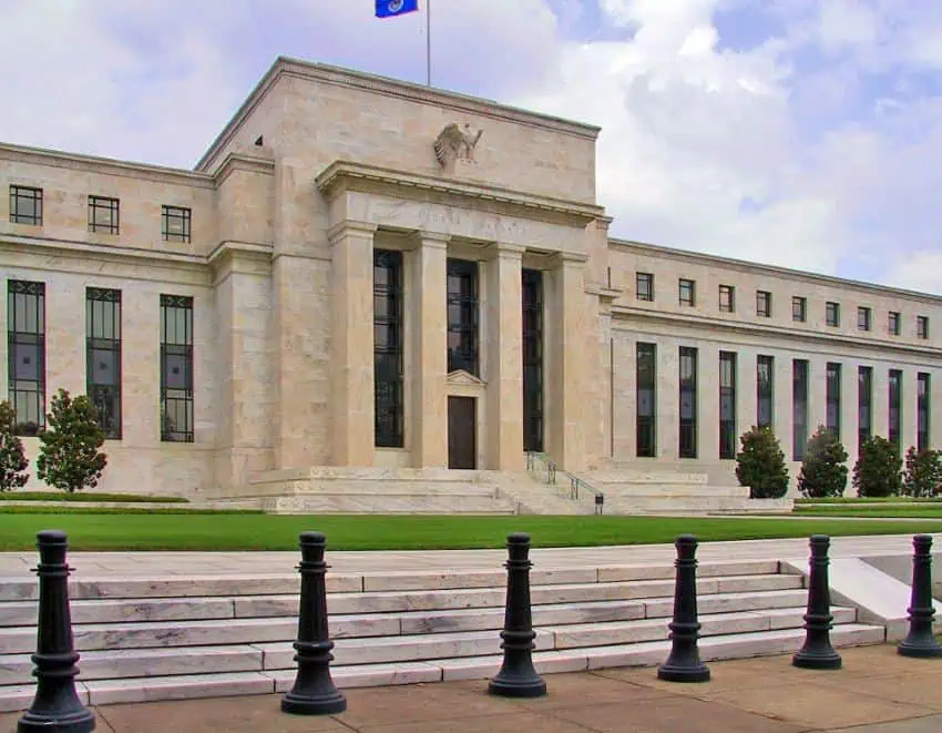 The U.S. Federal Reserve building, a wide marble building in Classical architectural style with several tall thin windows in the ground floor of the facade. It has a close cropped green lawn in front and a wide set of stairs that lead down into a courtyard