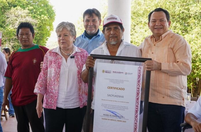 Mexico's Environment Minister, Alicia Barcena, a middle-aged woman with graying hair stands next to an unidentified Mexican man holding a framed enlarged document of certification of Sacpukenha as one of three new Maya protected areas. Around them are other people smiling into the camera.