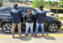 Two police officers with their backs to the camera standing in front of a police sedan. Their jackets say "SSPC Investigacion." Between them is a man facing the camera with his arms cuffed behind his back. His eyes are digitally covered with a black bar to hide his identity.