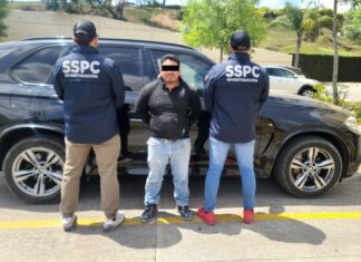 Two police officers with their backs to the camera standing in front of a police sedan. Their jackets say "SSPC Investigacion." Between them is a man facing the camera with his arms cuffed behind his back. His eyes are digitally covered with a black bar to hide his identity.