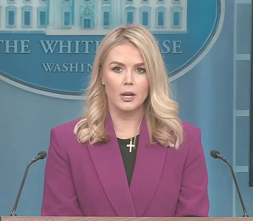 U.S. White House Press Secretary Karoline Leavitt standing at a podium with two microphones on either side of her and the White House logo on the wall behind her as she talks to news reporters in the White House Press Briefing Room.