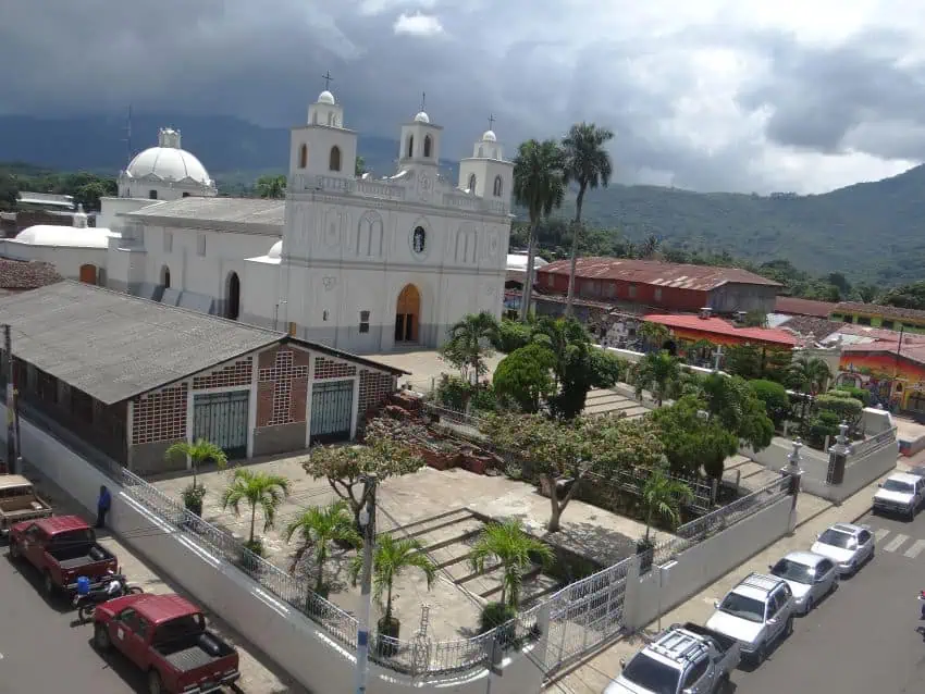 Aerial view of downtown Ahuachapán, El Salvador. Lines of cars are parked along the sidewalk bordering a church courtyard on a street corner.