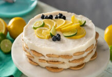 A round Carlota de limon cake with white cream cheese and condensed milk frosting with layers of round cookies presented on a white cake plate. It is topped with slices of lemon, lime and blueberries