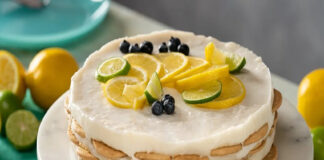 A round Carlota de limon cake with white cream cheese and condensed milk frosting with layers of round cookies presented on a white cake plate. It is topped with slices of lemon, lime and blueberries