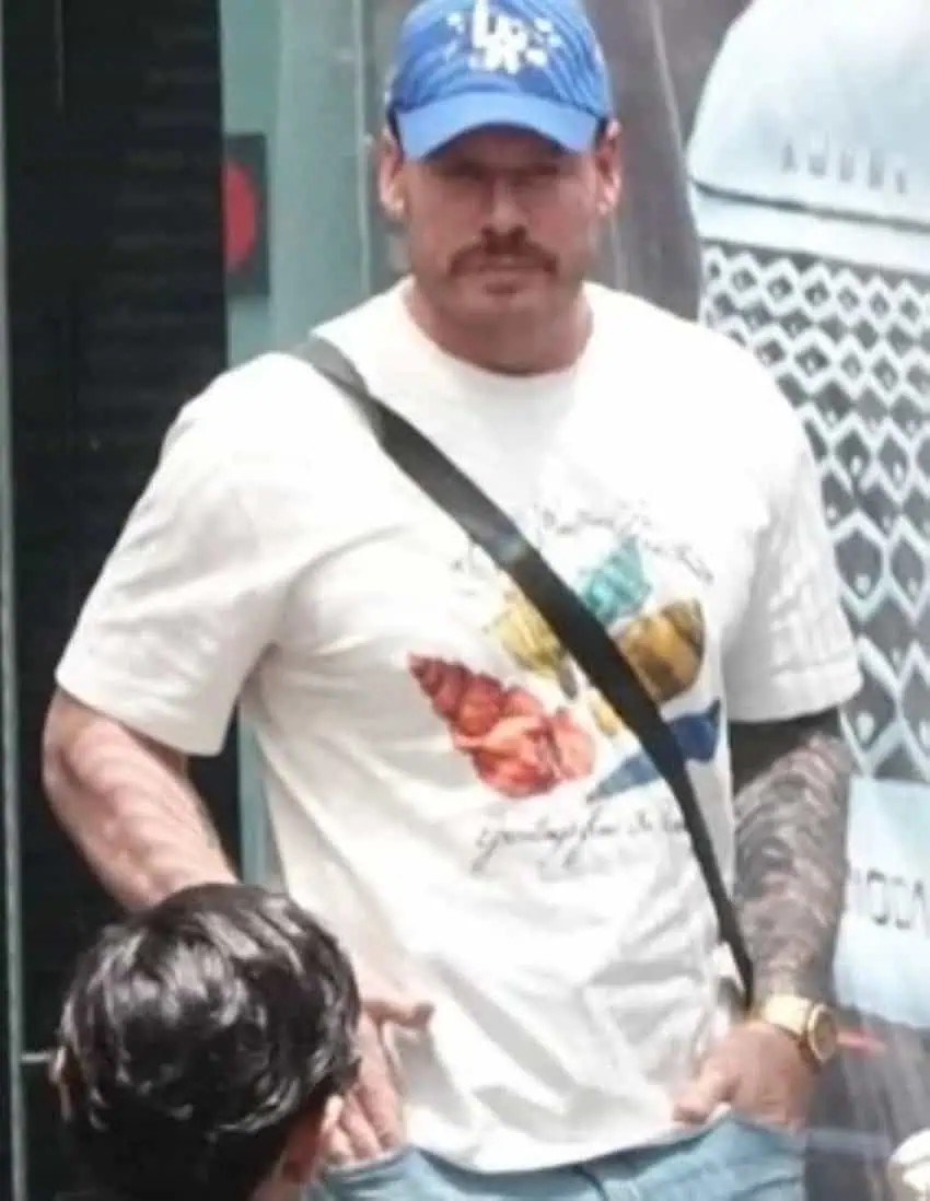 A middle-aged man with a mustache wearing a white teeshirt and a blue baseball cap stares into the camera as someone stands in the foreground left corner looking at him.