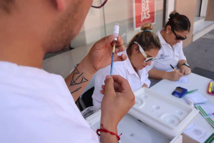 Three medical personnel in white lab coats, two sitting at a table, one standing in front of them in the foreground extracting a vaccine from a vial. The people at the table are reading and writing paperwork related to vaccinations.