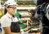 A young female Mexican worker wearing a factory-floor apron and hardhat as she uses a high-powered screwdriver on the steering wheel colum of a half-assembled vehicle