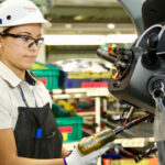 A young female Mexican worker wearing a factory-floor apron and hardhat as she uses a high-powered screwdriver on the steering wheel colum of a half-assembled vehicle