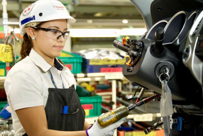 A young female Mexican worker wearing a factory-floor apron and hardhat as she uses a high-powered screwdriver on the steering wheel colum of a half-assembled vehicle