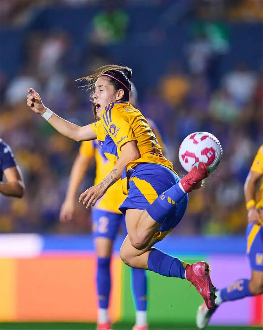 A woman soccer player in a blue and yellow uniform does something like a scorpion kick, hitting the ball with bottom of her cleat while flying through the air