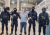 Guatemala's most wanted fugitive, La Chicharra, stands in a Guatemalan airport wearing a blue T-shirt, surrounded by masked soldiers in front of a sign reading "Welcome to Guatemala"