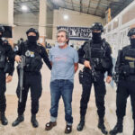 Guatemala's most wanted fugitive, La Chicharra, stands in a Guatemalan airport wearing a blue T-shirt, surrounded by masked soldiers in front of a sign reading "Welcome to Guatemala"