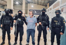 Guatemala's most wanted fugitive, La Chicharra, stands in a Guatemalan airport wearing a blue T-shirt, surrounded by masked soldiers in front of a sign reading "Welcome to Guatemala"