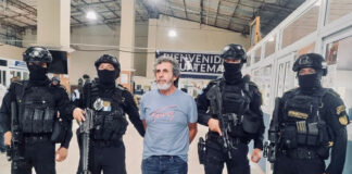 Guatemala's most wanted fugitive, La Chicharra, stands in a Guatemalan airport wearing a blue T-shirt, surrounded by masked soldiers in front of a sign reading "Welcome to Guatemala"