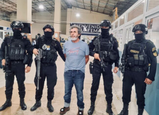 Guatemala's most wanted fugitive, La Chicharra, stands in a Guatemalan airport wearing a blue T-shirt, surrounded by masked soldiers in front of a sign reading "Welcome to Guatemala"