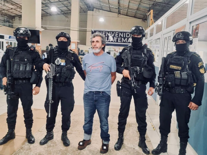 Guatemala's most wanted fugitive, La Chicharra, stands in a Guatemalan airport wearing a blue T-shirt, surrounded by masked soldiers in front of a sign reading 