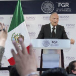 Attorney General Alejandro Gertz Manero stands at a podium giving a report on the Jalisco extermination camp case, next to a Mexican flag as reporters raise their hands in the foreground