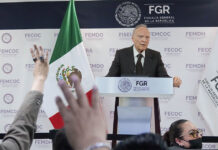 Attorney General Alejandro Gertz Manero stands at a podium giving a report on the Jalisco extermination camp case, next to a Mexican flag as reporters raise their hands in the foreground